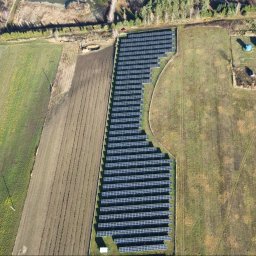 EKO ENERGIA KRZYSZTOF SZPYRA - Widok z lotu ptaka na rozległą farmę fotowoltaiczną, otoczoną polami uprawnymi i lasem, z budynkami w tle. Panele ułożone rzędami, tworzą geometryczny wzór na zielonym terenie.