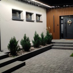 EKO ENERGIA KRZYSZTOF SZPYRA - Eleganckie oświetlenie LED podkreśla nowoczesną fasadę domu z drewnianymi panelami i stylowymi schodami wejściowymi, tworząc przytulną atmosferę.