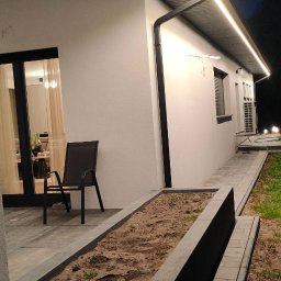 EKO ENERGIA KRZYSZTOF SZPYRA - Nowoczesny dom z oświetleniem zewnętrznym LED pod dachem, oświetlona ścieżka i ogród. Widok nocny, z boku domu, z krzesłem na tarasie.