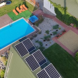 EKO ENERGIA KRZYSZTOF SZPYRA - Widok z góry na dom z basenem i panelami słonecznymi na dachu. Na leżakach obok basenu leżą dwie osoby. W tle widoczny ogród z trampoliną i huśtawką.