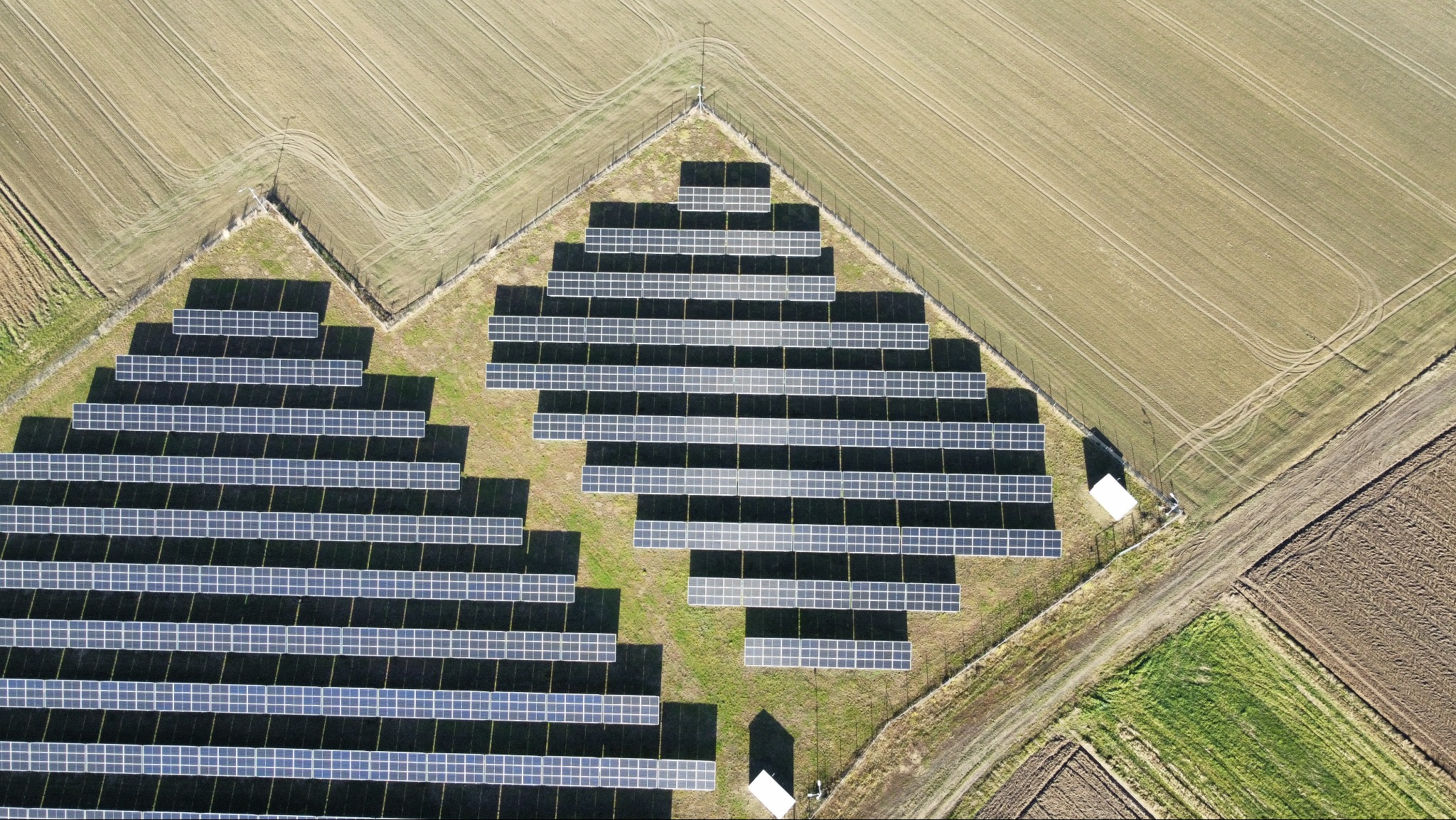 Widok z lotu ptaka na rozległą farmę fotowoltaiczną, otoczoną polami uprawnymi o różnych odcieniach brązu i zieleni. Panele ułożone w rzędach, tworzą geometryczny wzór.