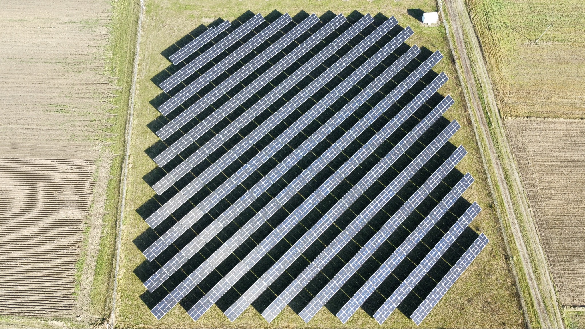 Widok z góry na dużą farmę fotowoltaiczną z panelami ułożonymi rzędami na zielonym terenie, obok pola uprawnego i drogi polnej. Biały budynek w tle.