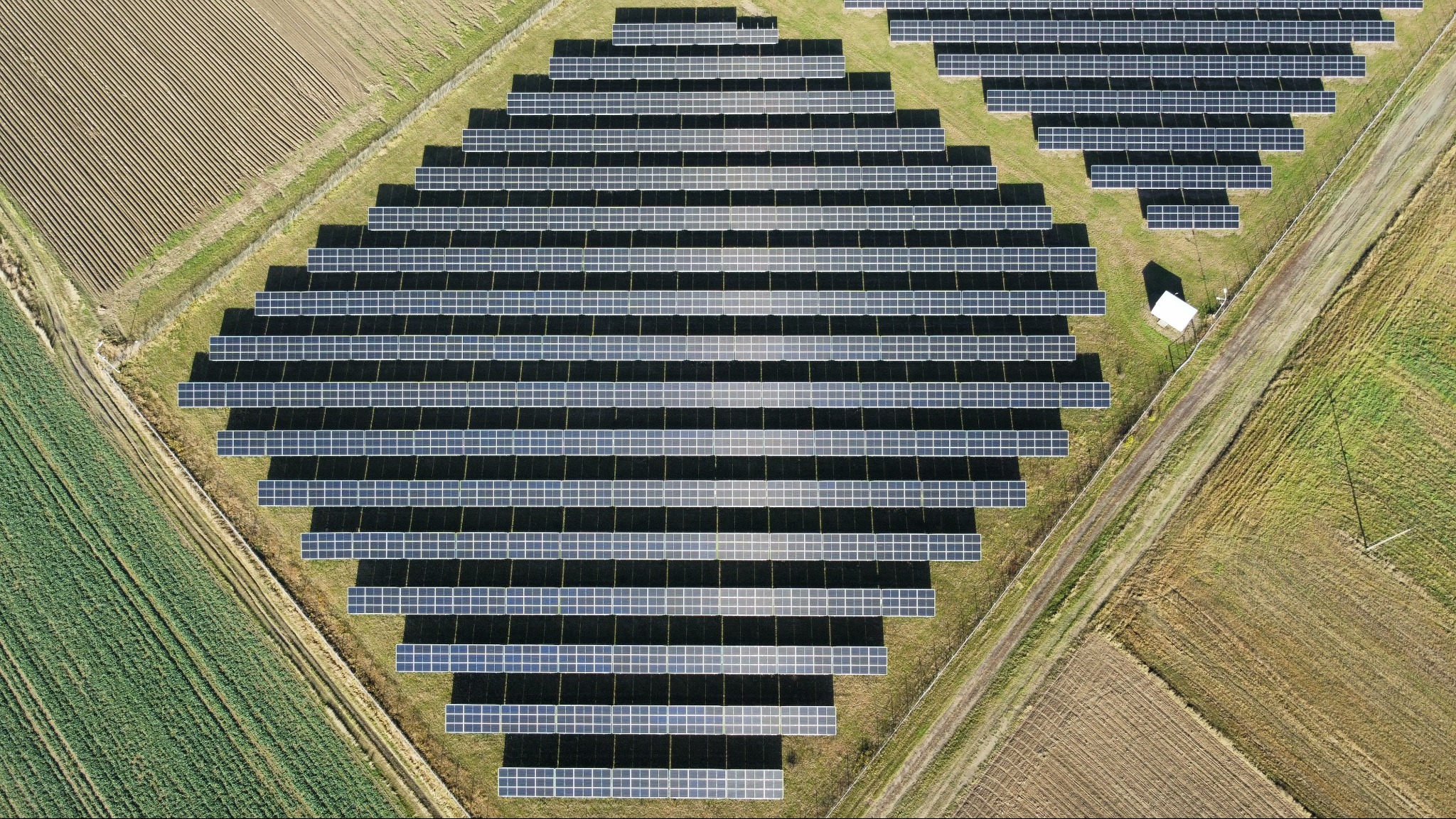 Widok z lotu ptaka na dużą farmę fotowoltaiczną w kształcie rombu, otoczoną polami uprawnymi i małym budynkiem technicznym. Panele ułożone w rzędach.