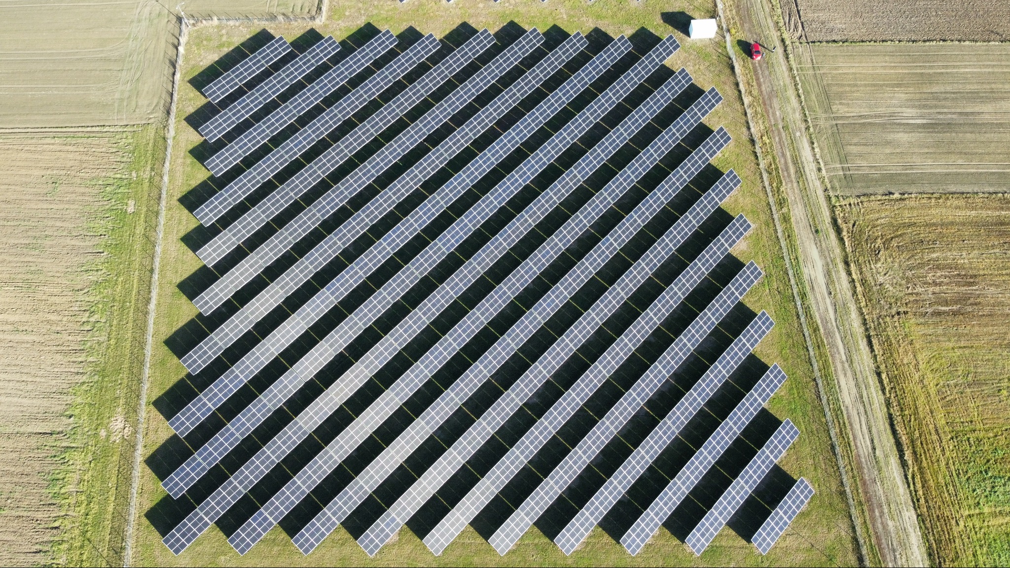 Widok z drona na dużą farmę fotowoltaiczną z panelami ułożonymi rzędami na zielonym polu, obok pola uprawne i mały budynek gospodarczy z czerwonym autem.