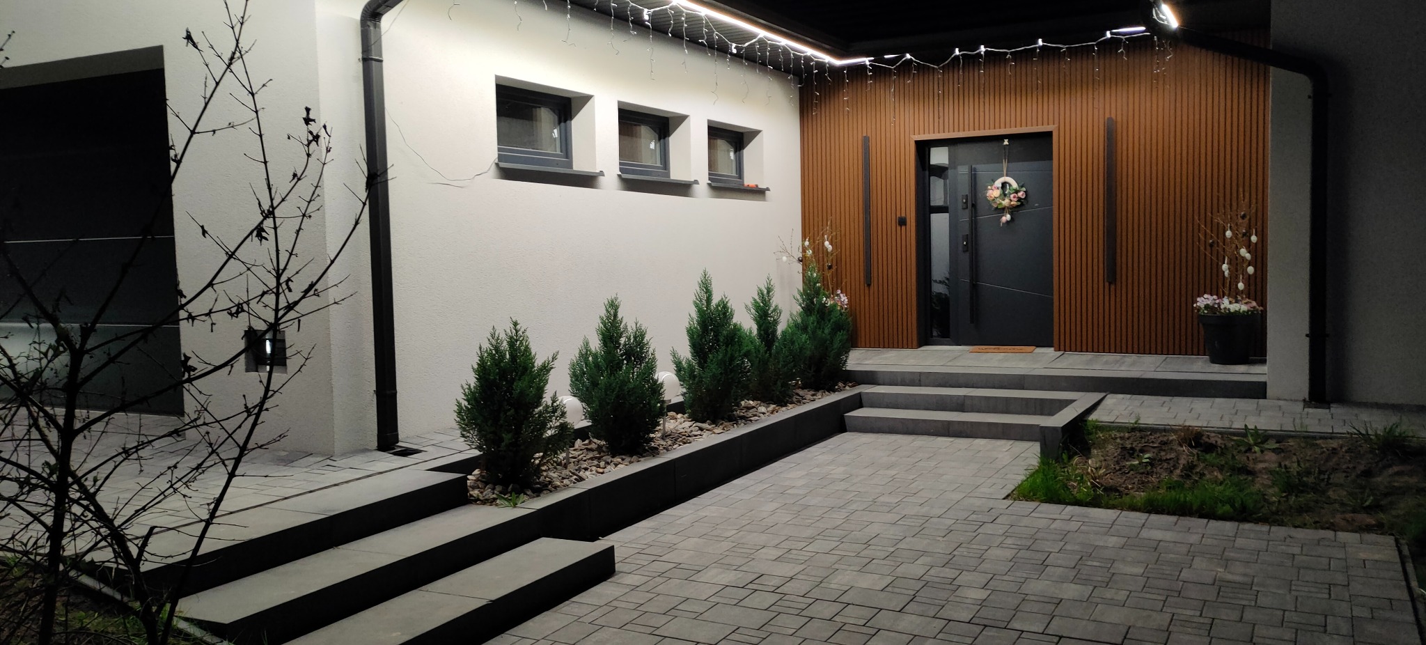 Eleganckie oświetlenie LED podkreśla nowoczesną fasadę domu z drewnianymi panelami i stylowymi schodami wejściowymi, tworząc przytulną atmosferę.