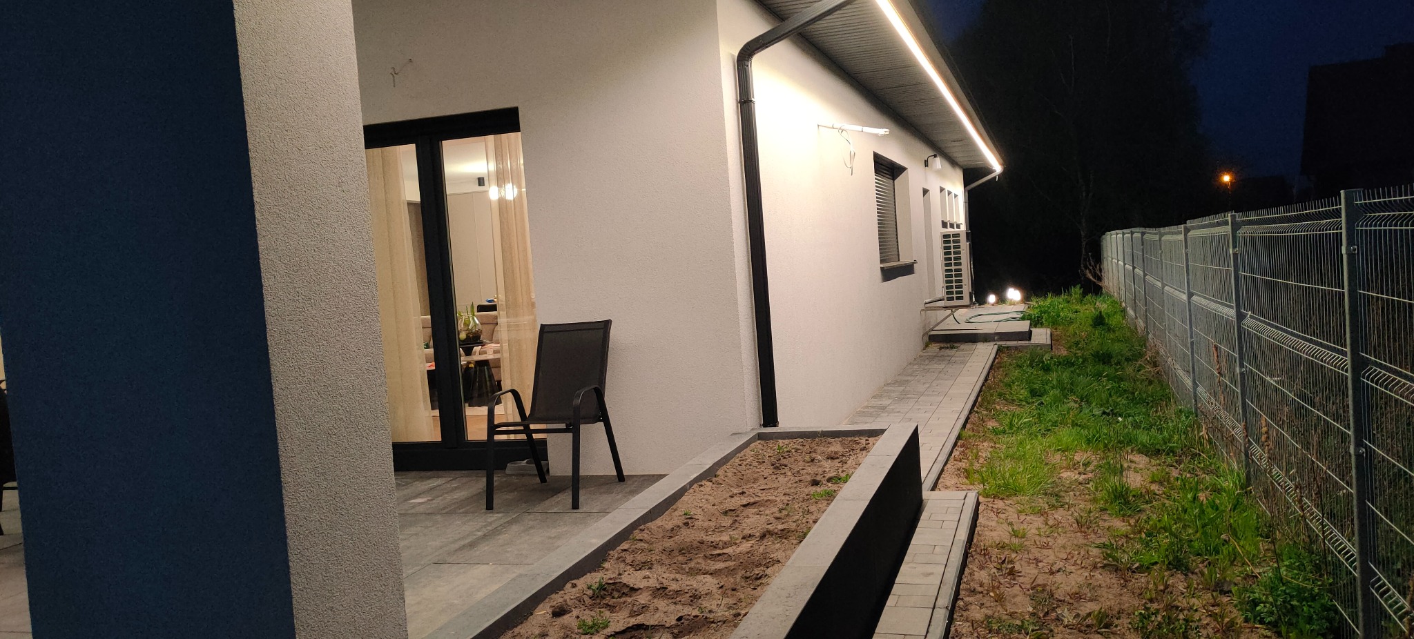 Nowoczesny dom z oświetleniem zewnętrznym LED pod dachem, oświetlona ścieżka i ogród. Widok nocny, z boku domu, z krzesłem na tarasie.