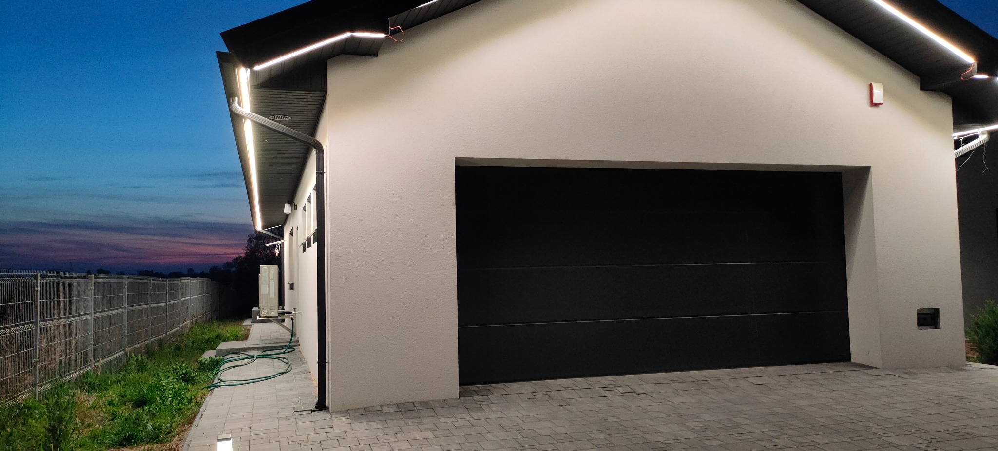 Nowoczesny dom z garażem i oświetleniem LED na elewacji, widok z zewnątrz o zmierzchu. Czarna brama garażowa, szara kostka brukowa i fragment ogrodzenia.