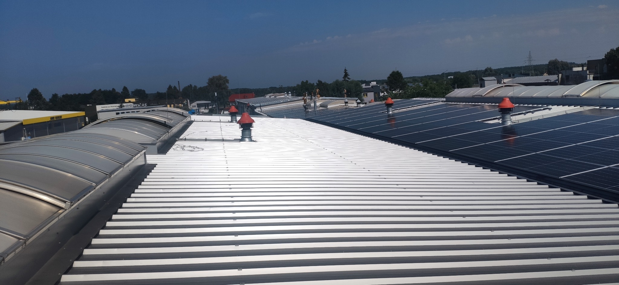 Instalacja fotowoltaiczna na dachu hali z blachy trapezowej, widoczne panele słoneczne i wentylatory dachowe z czerwonymi daszkami. Ujęcie z góry, w tle zabudowania.
