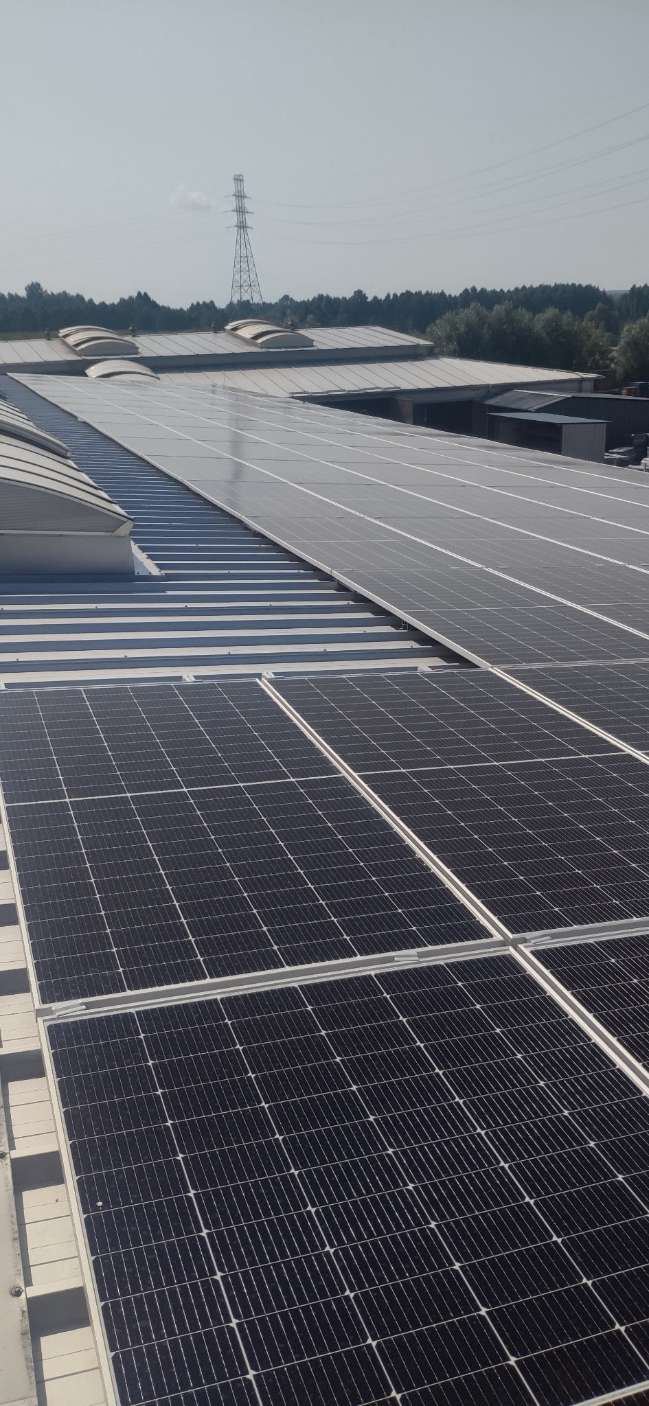 Rząd paneli fotowoltaicznych na dachu budynku przemysłowego, w tle słup wysokiego napięcia i zieleń. Widok z góry, oświetlenie dzienne.