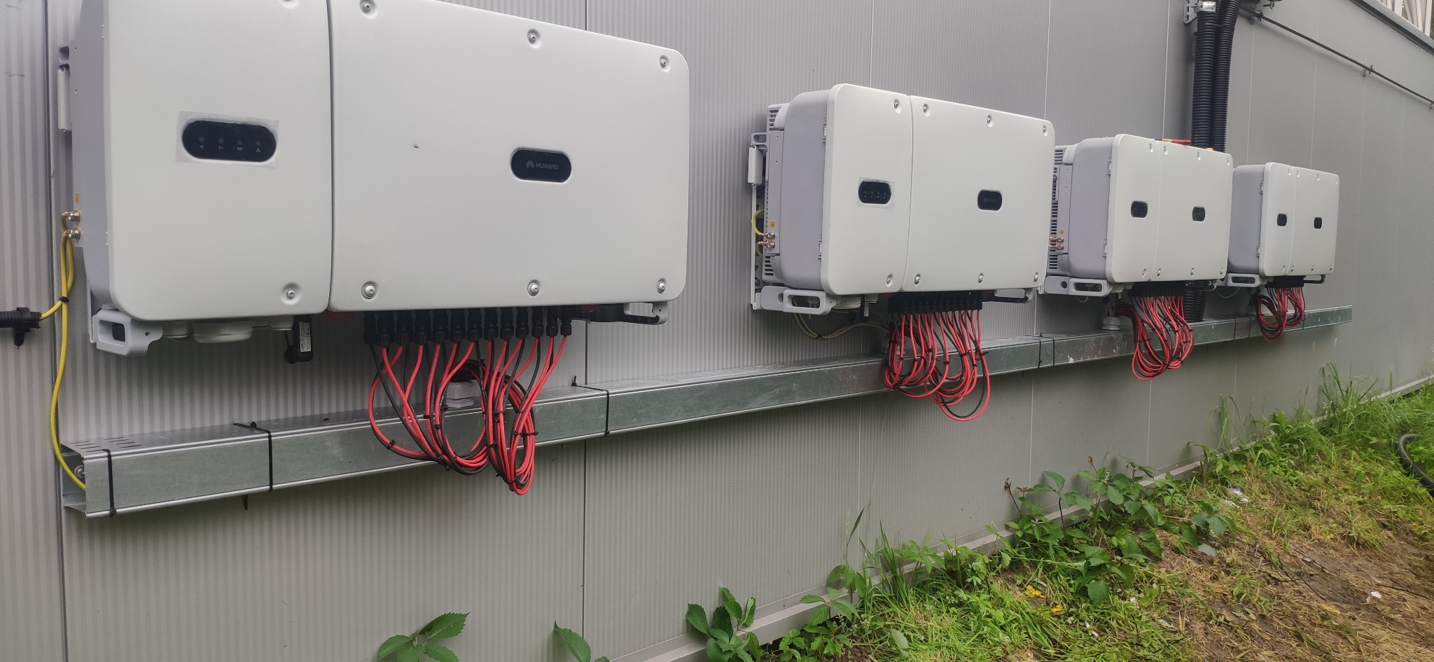 Rząd falowników fotowoltaicznych Huawei zamontowanych na metalowej szynie przy ścianie budynku w Płoskiem. Widoczne kable i złącza.