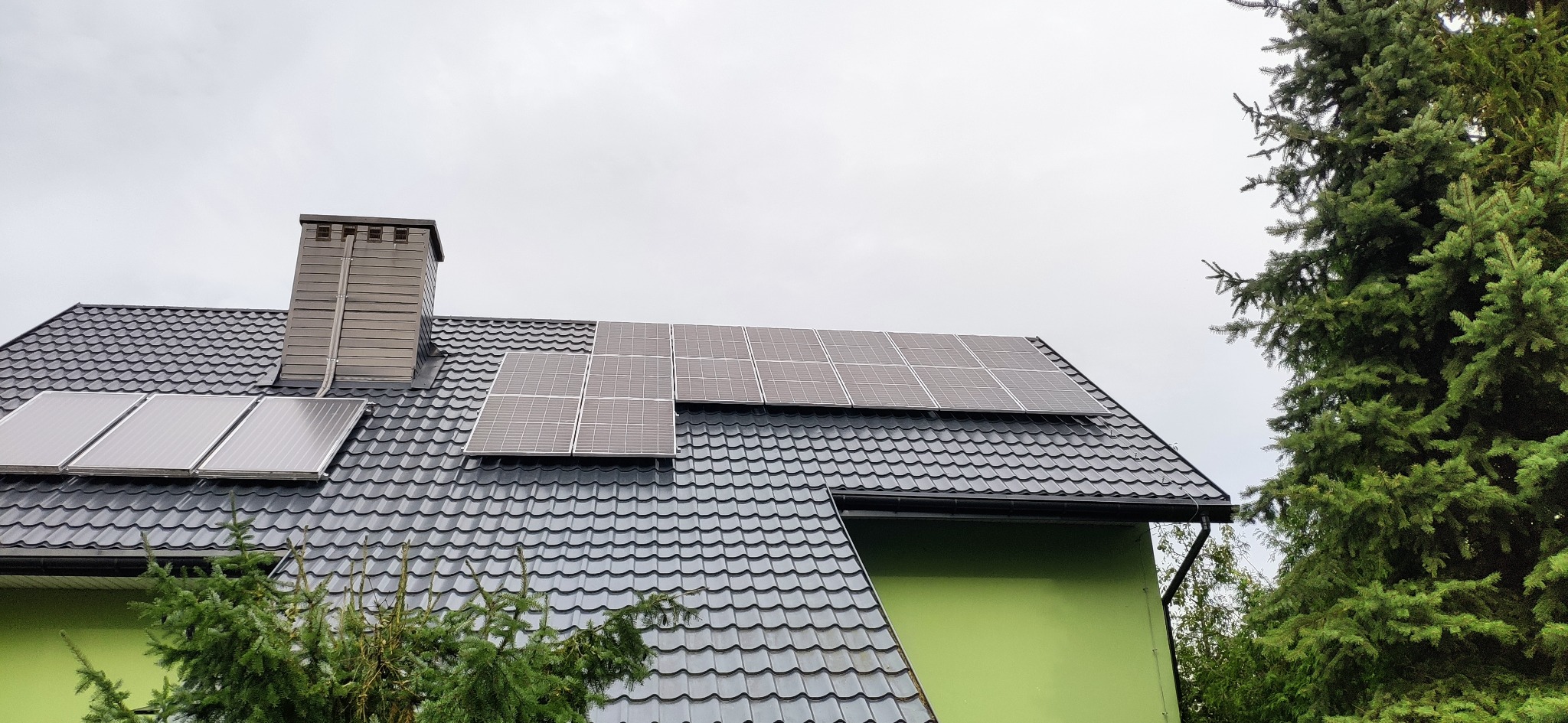 Panele fotowoltaiczne na dachu domu z szarej blachy, komin z brązowymi panelami, zielona ściana i drzewo iglaste. Pochmurne niebo w tle.