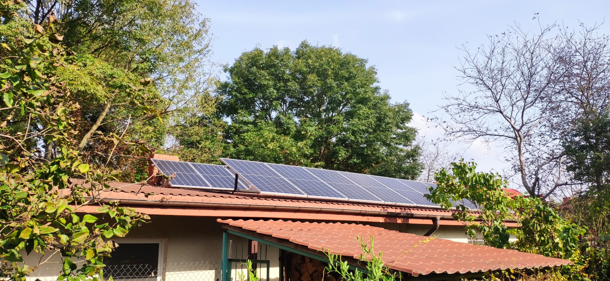 Panele fotowoltaiczne na dachu domu jednorodzinnego, widoczne z poziomu gruntu, otoczone zielenią drzew. Czerwona dachówka i błękitne niebo w tle.