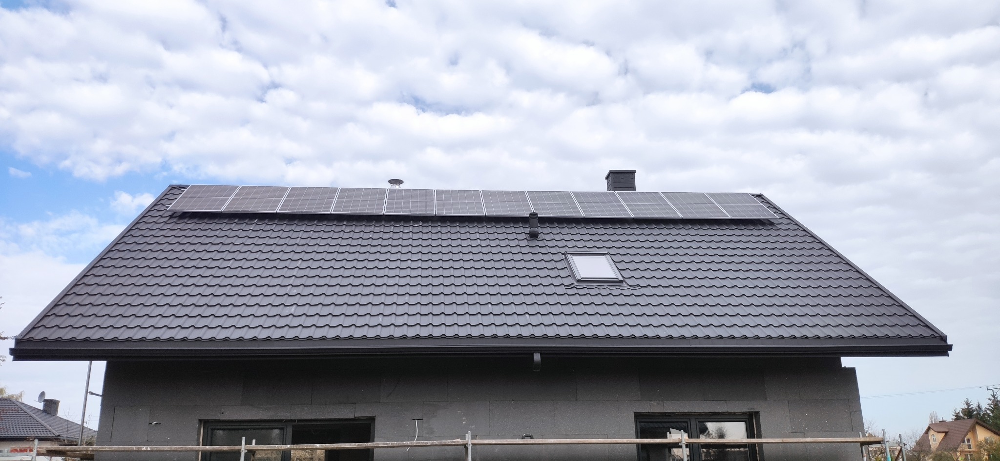 Czarny dach z panelami fotowoltaicznymi i oknem dachowym na tle pochmurnego nieba. Dom w trakcie budowy, widoczne rusztowania.