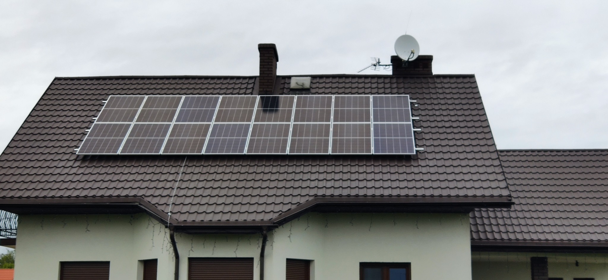 Brązowy dach z panelami słonecznymi, antena satelitarna i komin. Widok na dom z zewnątrz w pochmurny dzień. Fotowoltaika na dachu domu jednorodzinnego.