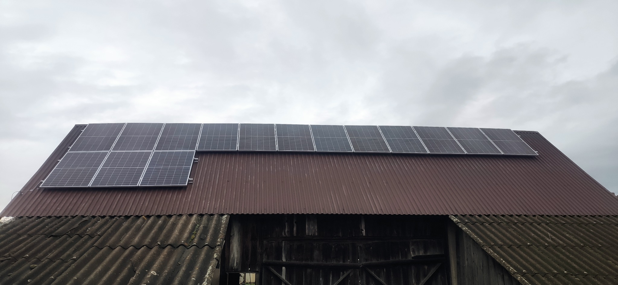 Panele fotowoltaiczne na dachu starej, brązowej stodoły pod pochmurnym niebem. Widok z dołu, ukazujący instalację na tle wiejskiego krajobrazu.