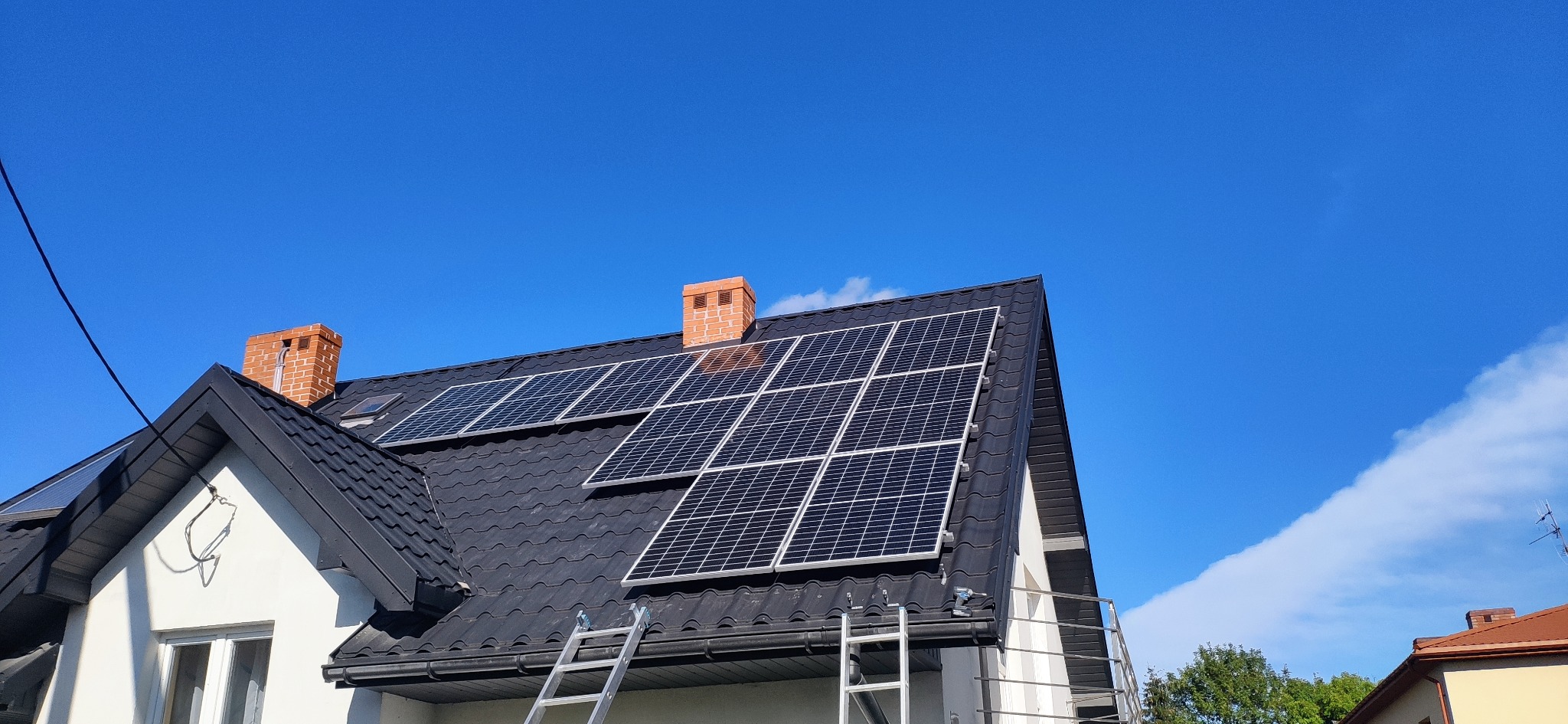 Panele fotowoltaiczne na dachu domu jednorodzinnego z czarną dachówką, drabina oparta o dach, błękitne niebo w tle. Widok z dołu.