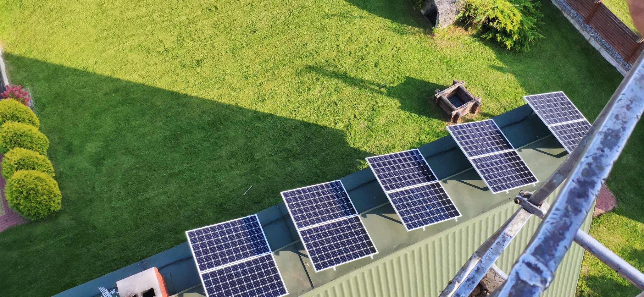 Widok z góry na panele fotowoltaiczne na zielonym dachu, z fragmentem trawnika i elementami konstrukcji wsporczej w kadrze. Ujęcie z perspektywy drabiny.