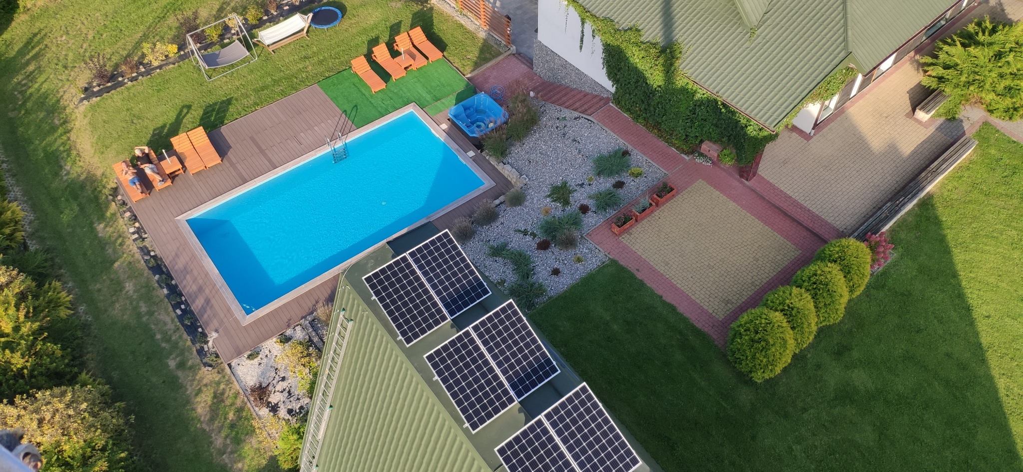 Widok z góry na dom z basenem i panelami słonecznymi na dachu. Na leżakach obok basenu leżą dwie osoby. W tle widoczny ogród z trampoliną i huśtawką.