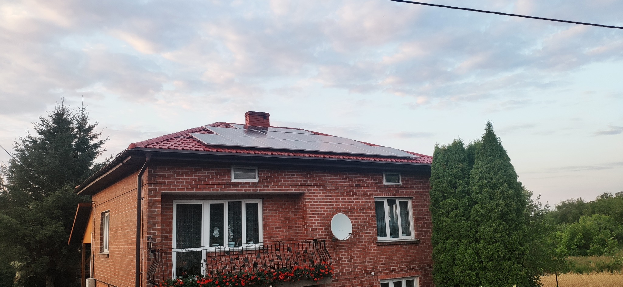 Ceglany dom z czerwoną dachówką i panelami fotowoltaicznymi na dachu, balkon z kwiatami, drzewa w tle, pochmurne niebo.