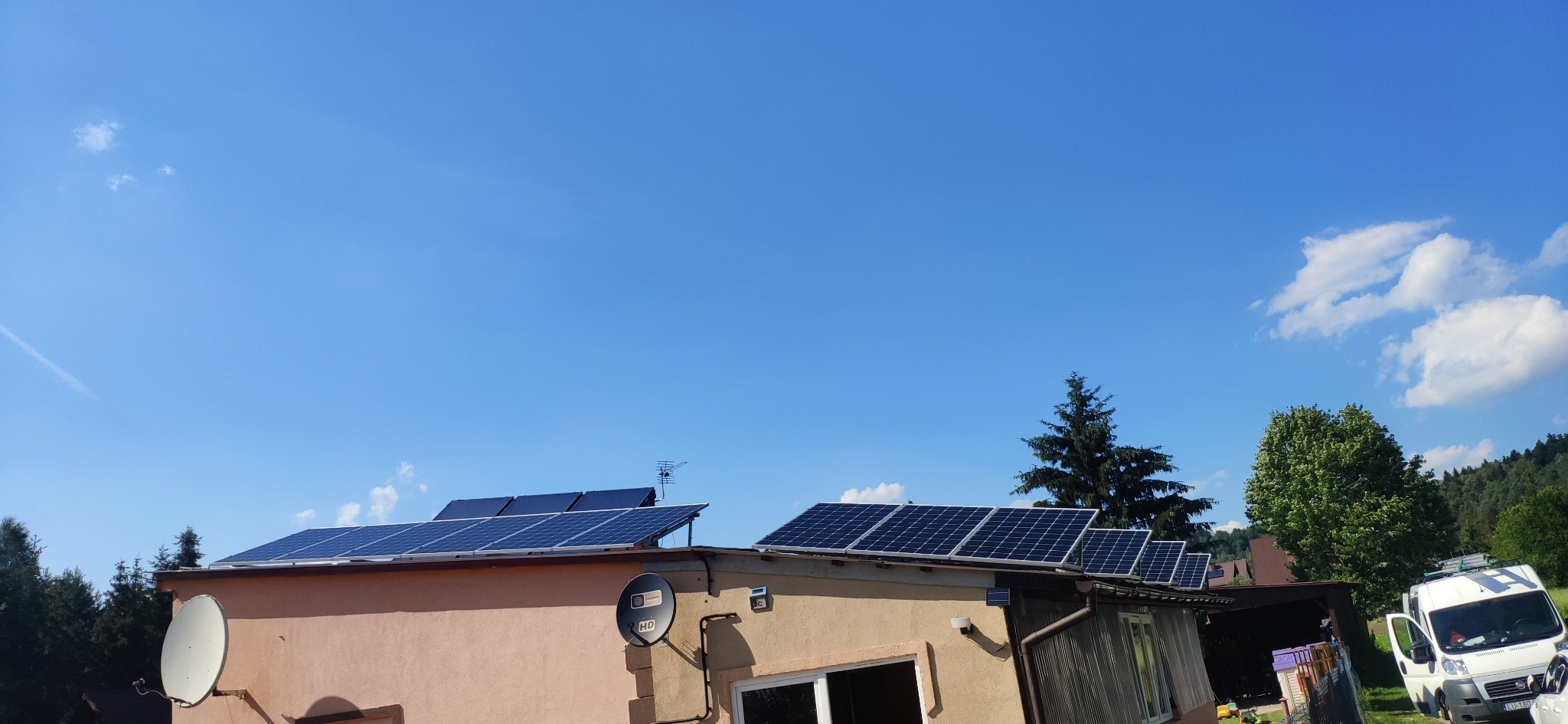 Panele fotowoltaiczne na dachu budynku z anteną satelitarną, na tle błękitnego nieba z chmurami. W tle drzewa i częściowo widoczny samochód dostawczy.