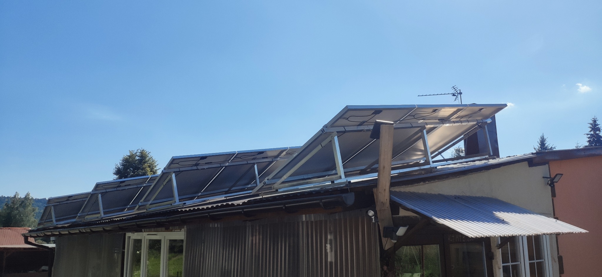 Panele fotowoltaiczne zamontowane na dachu budynku z blachy falistej, widoczne mocowania i okablowanie, błękitne niebo w tle, antena telewizyjna na dachu.