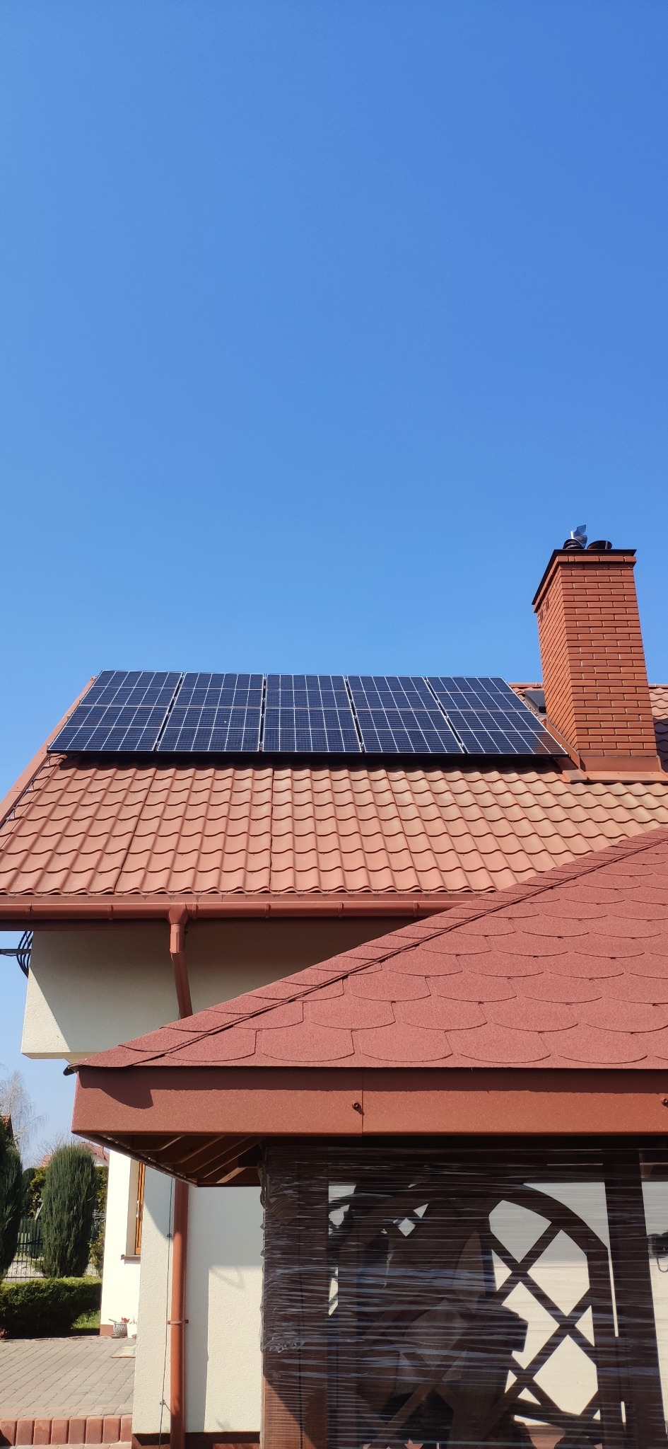 Panele fotowoltaiczne na dachu domu z czerwoną dachówką i ceglanym kominem, błękitne niebo w tle. Widoczny fragment altany z folią ochronną.