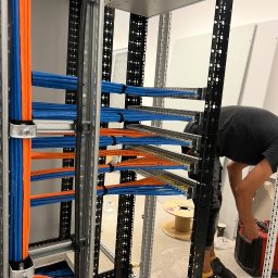 DAWID WÓJCIAK - Uporządkowane kable w kolorach niebieskim i pomarańczowym, ułożone w metalowej konstrukcji szafy serwerowej. Widoczny fragment osoby pracującej nad instalacją.