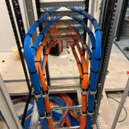 Profesjonalnie ułożone kable w szafie serwerowej, uporządkowane w wiązki niebieskie i pomarańczowe, zabezpieczone czarnymi opaskami zaciskowymi. Widoczne elementy konstrukcji szafy.