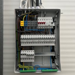 DAWID WÓJCIAK - Otwarta skrzynka elektryczna z bezpiecznikami, widoczne kolorowe przewody i złączki. Instalacja zamontowana na białej, panelowej ścianie.