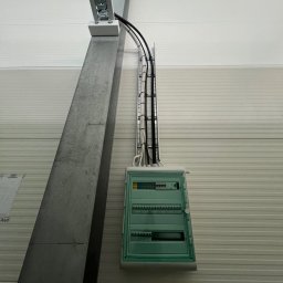 DAWID WÓJCIAK - Zielona skrzynka elektryczna z kablami zamontowana na ścianie z blachy trapezowej, widoczny metalowy słup i koryto kablowe. Ujęcie z dołu, oświetlenie górne.