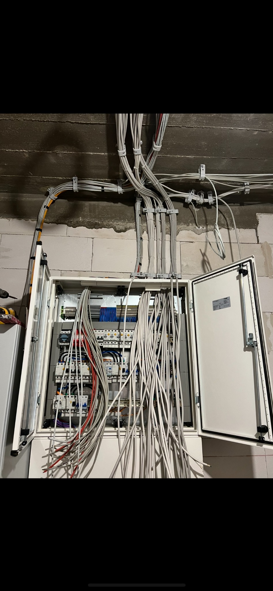 Otwarta skrzynka elektryczna z licznymi przewodami wewnątrz i na zewnątrz, zamocowanymi do sufitu w surowym wnętrzu. Widoczne bezpieczniki i inne elementy instalacji.