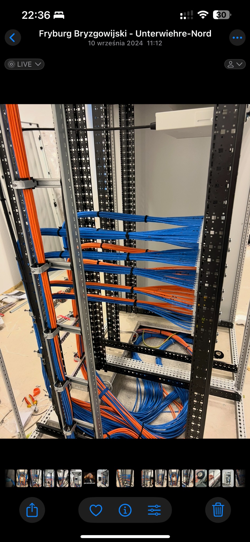 Instalacja okablowania strukturalnego: pomarańczowe i niebieskie przewody uporządkowane w metalowej szafie rack, widoczne narzędzia i elementy montażowe.