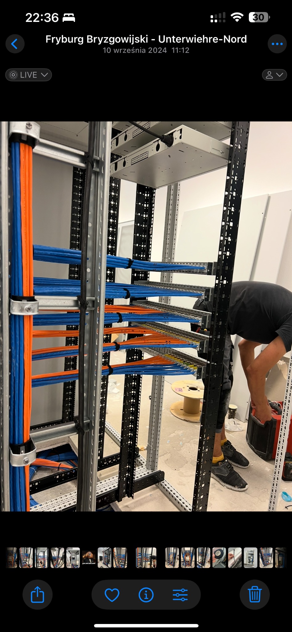 Uporządkowane kable w kolorach niebieskim i pomarańczowym, ułożone w metalowej konstrukcji szafy serwerowej. Widoczny fragment osoby pracującej nad instalacją.