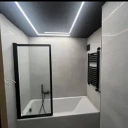 Wykończona łazienka z wanną i czarną armaturą. Kabina prysznicowa ze szkła, grzejnik drabinkowy. Białe płytki na ścianach, czarny sufit z ledowym oświetleniem.