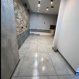 Perfectdrywall - Wykończone wnętrze: szare płytki podłogowe, ceglana ściana z elementami szarości, białe ściany i sufit z oświetleniem. Widoczny fragment wiadra.