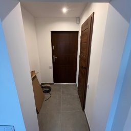 Perfectdrywall - Wnętrze korytarza po remoncie: szare płytki podłogowe, białe ściany, dwoje ciemnych drzwi. Widoczny karton i kable, sugerujące prace wykończeniowe.