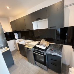 Perfectdrywall - Nowoczesna kuchnia w trakcie montażu: szare szafki wiszące i dolne, czarny panel nad blatem, sprzęt AGD częściowo zabezpieczony folią. Widoczne niedokończone prace.