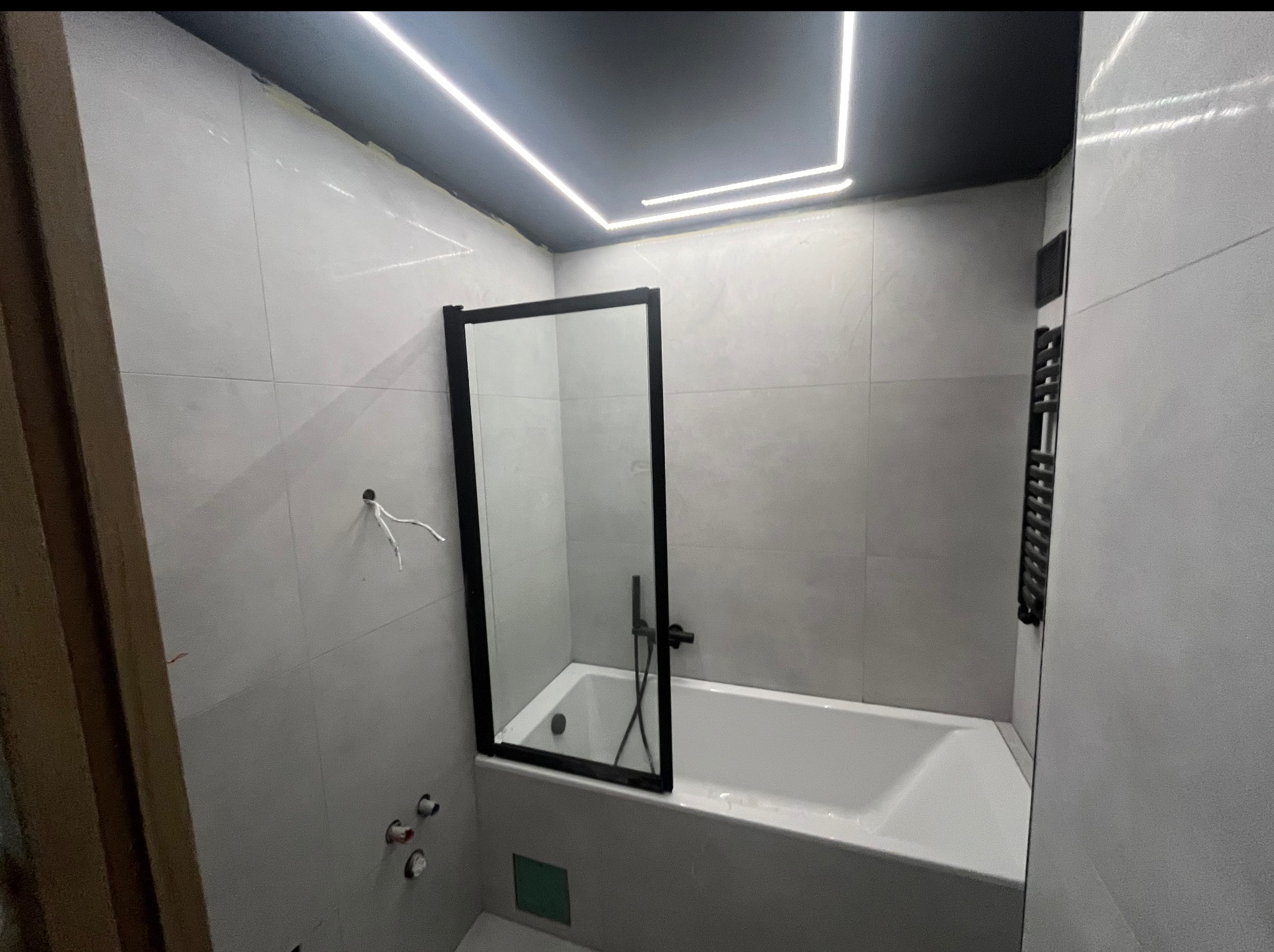 Nowoczesna łazienka po remoncie: białe płytki, czarna armatura, kabina nawannowa z czarną ramą i podświetlany sufit. Widoczne niedokończone elementy instalacji.