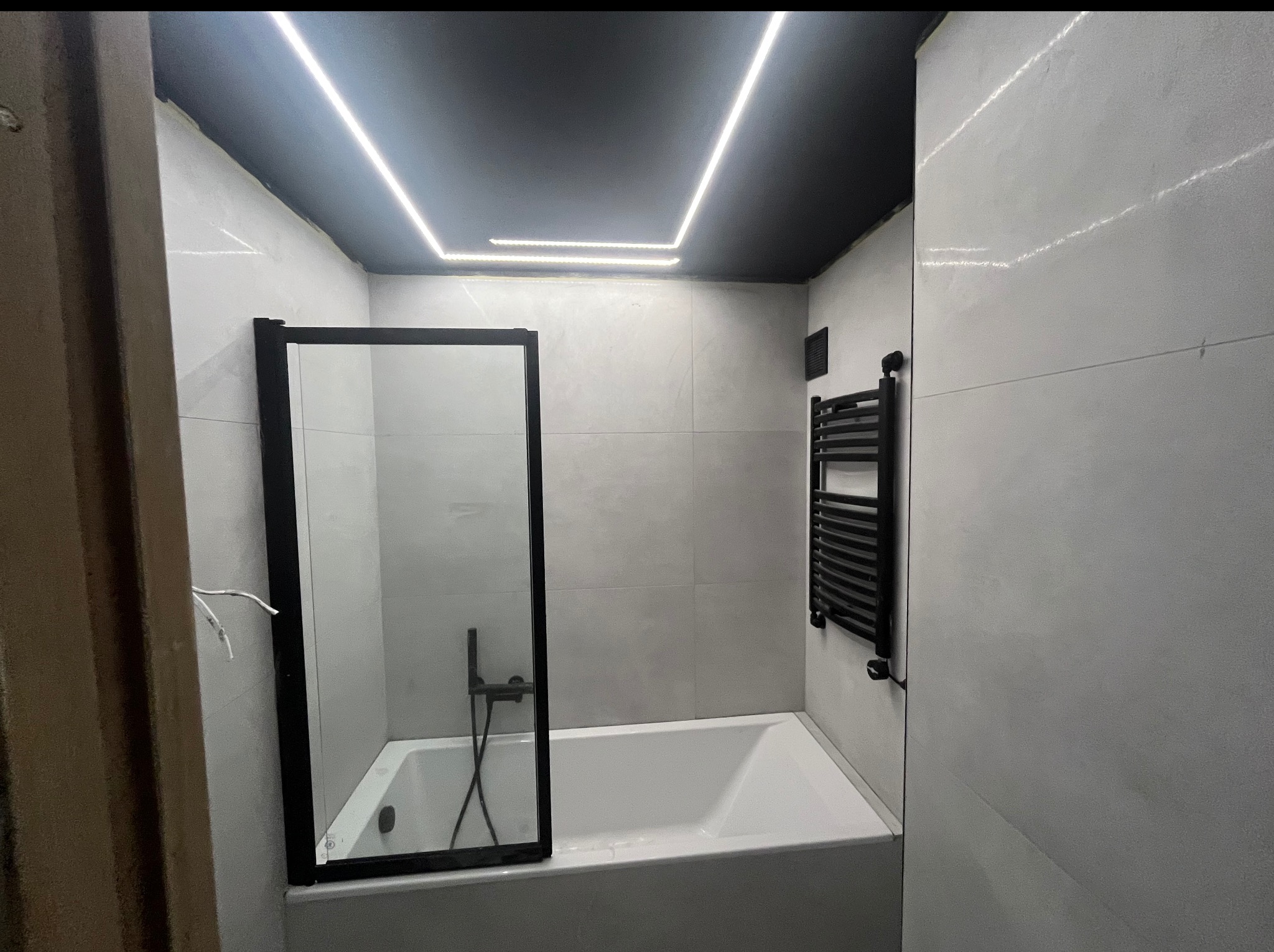 Wykończona łazienka z wanną i czarną armaturą. Kabina prysznicowa ze szkła, grzejnik drabinkowy. Białe płytki na ścianach, czarny sufit z ledowym oświetleniem.