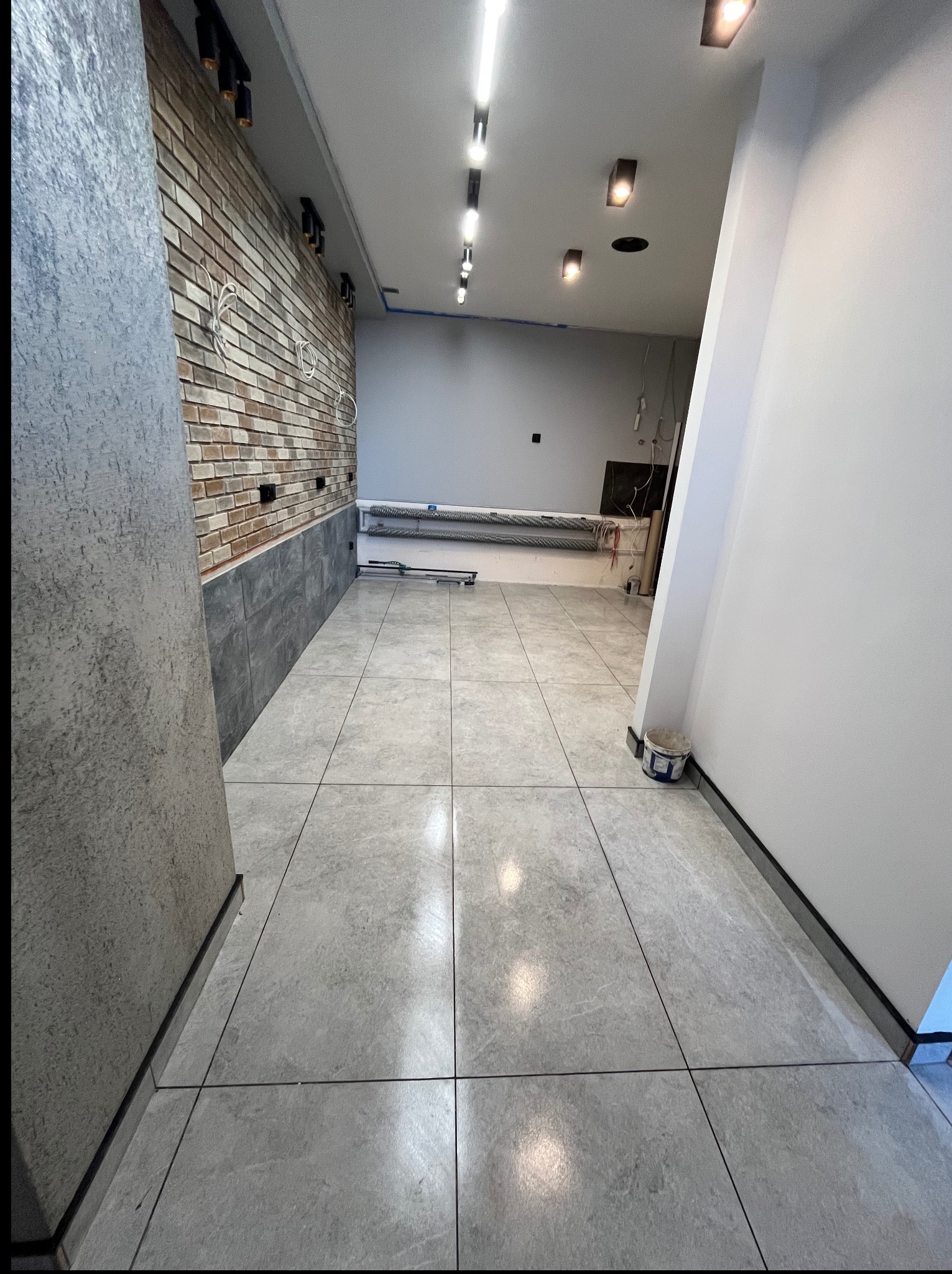 Wykończone wnętrze: szare płytki podłogowe, ceglana ściana z elementami szarości, białe ściany i sufit z oświetleniem. Widoczny fragment wiadra.