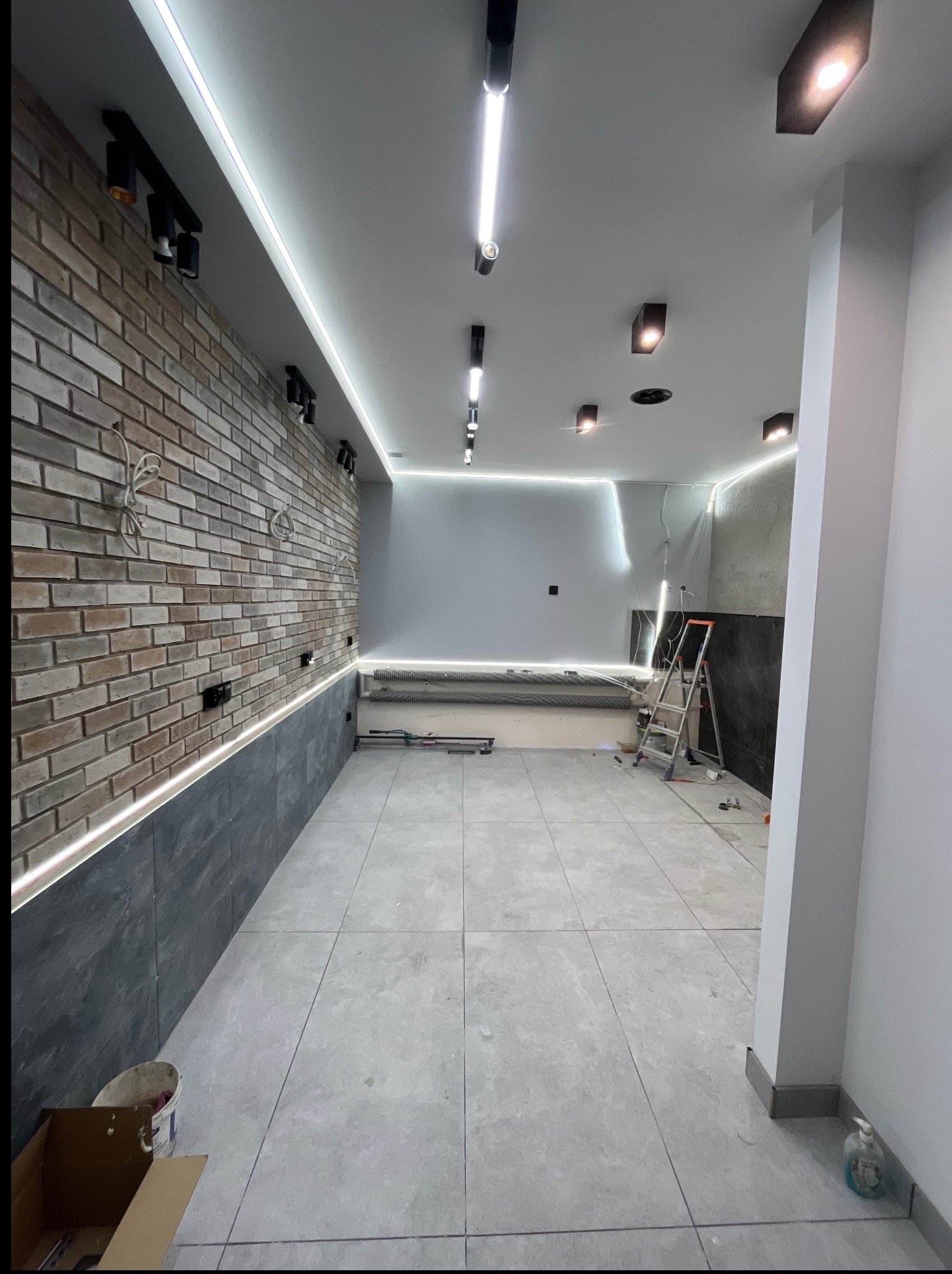 Wnętrze w trakcie wykańczania: ceglana ściana z oświetleniem LED, szare płytki na podłodze, drabina, surowy stan w tle, widoczne kable i puszki elektryczne.