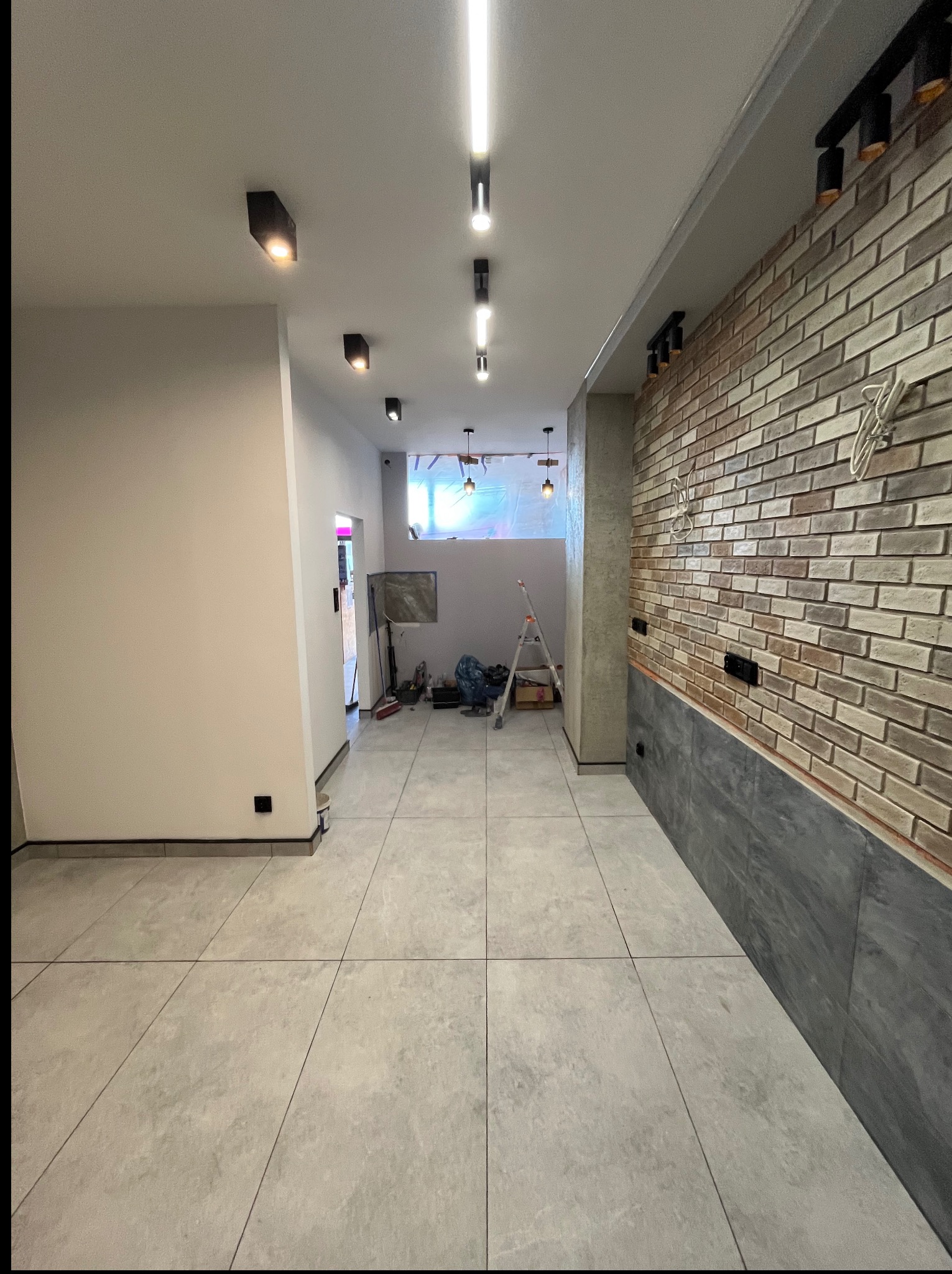 Wnętrze w trakcie wykańczania: szara podłoga, ściana z cegły, oświetlenie sufitowe. Widoczna drabina i narzędzia w tle, sugerujące trwające prace.