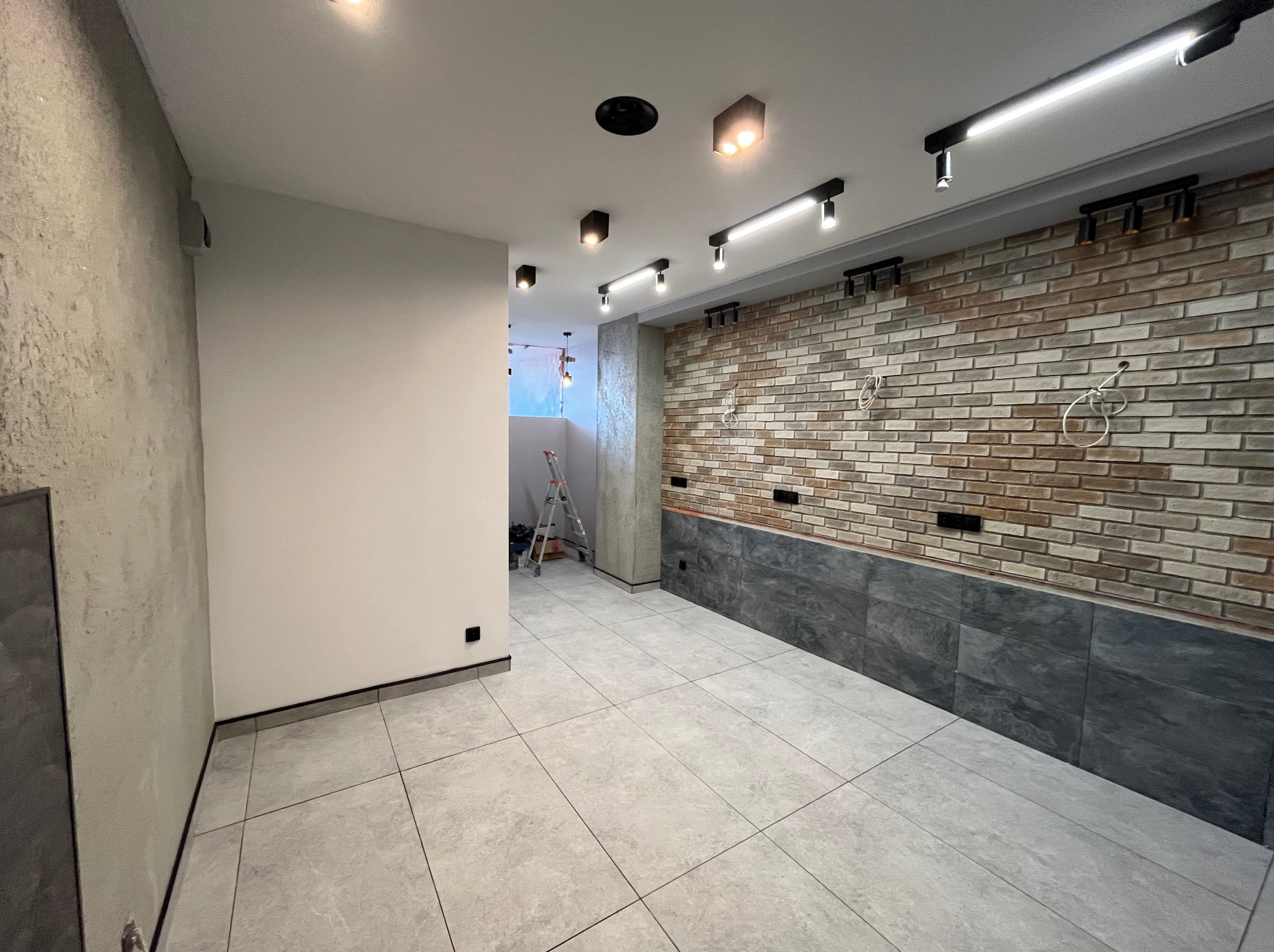 Wykończone wnętrze: szare płytki podłogowe, ściana z cegły i szara lamperia. Widoczne oświetlenie sufitowe i drabina w głębi pomieszczenia. Nowoczesny design.