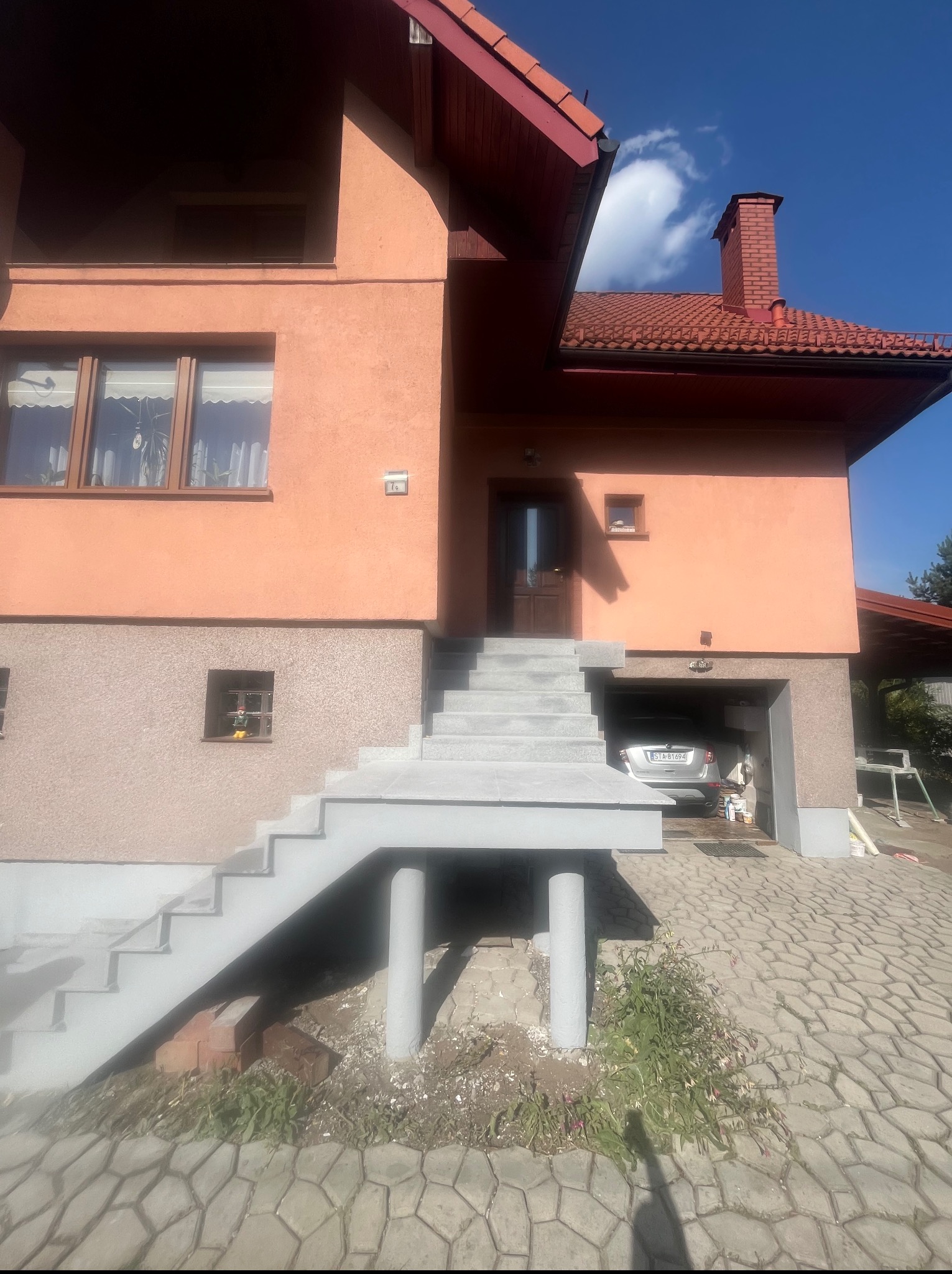 Dwukondygnacyjny dom z garażem, schodami wejściowymi podpartymi filarami i czerwoną dachówką. Na podjeździe kostka brukowa.