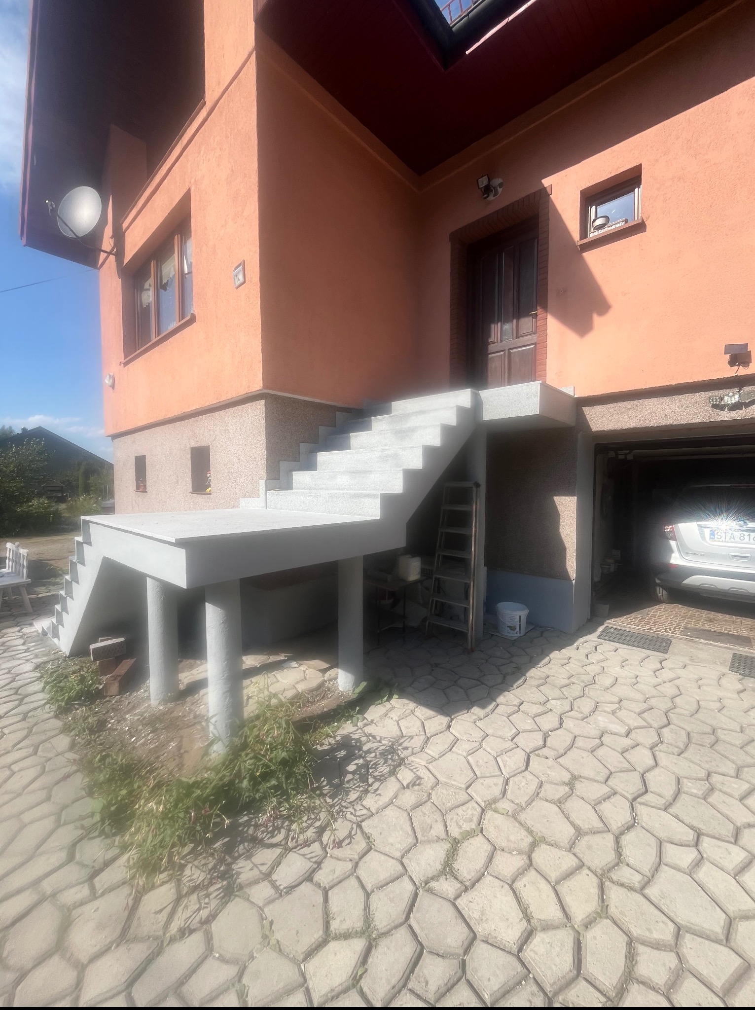 Nowe, betonowe schody zewnętrzne prowadzące do drzwi wejściowych domu z garażem. Widoczne narzędzia i materiały budowlane.