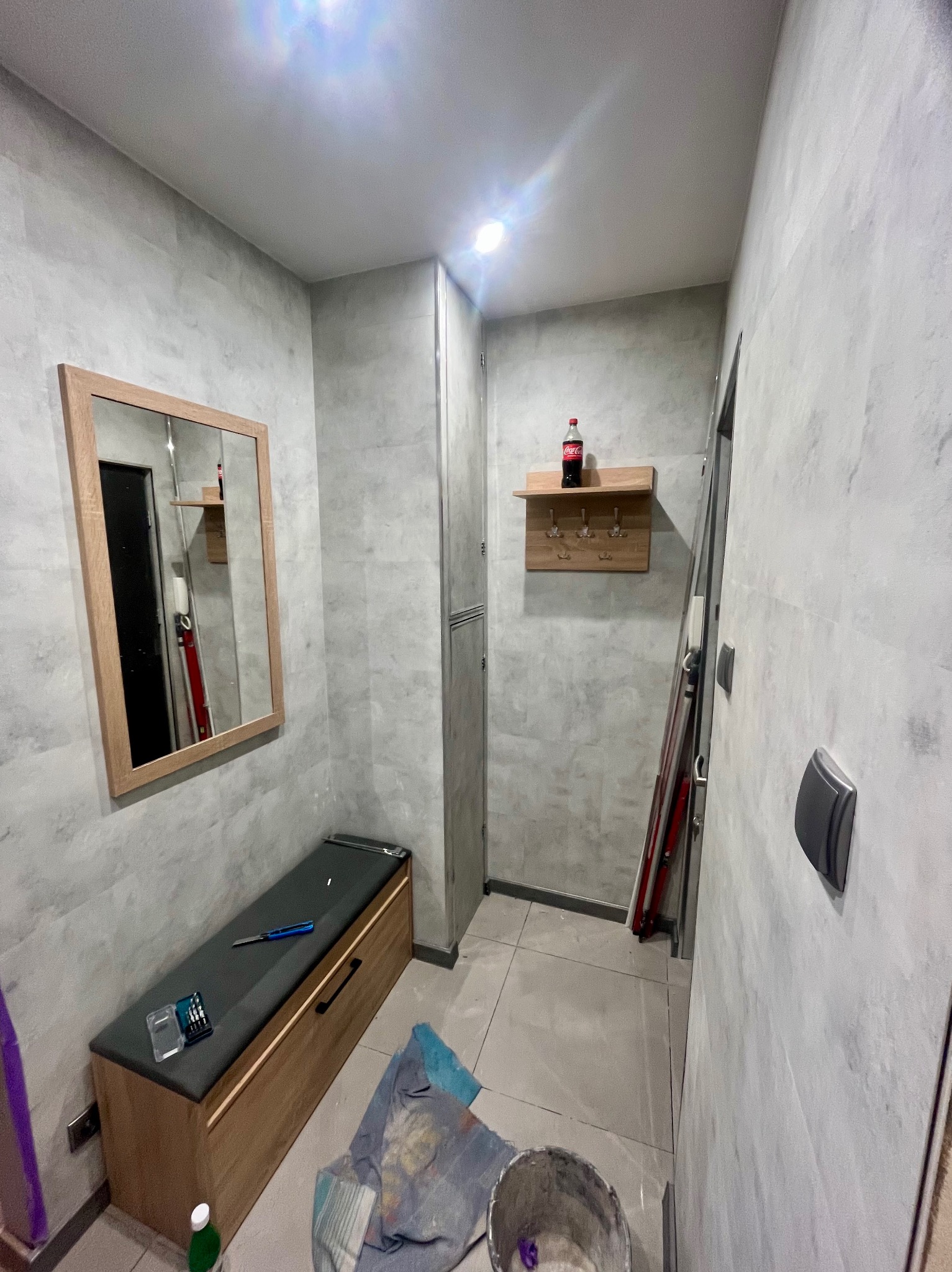 Wykończony korytarz w szarościach z lustrem w drewnianej ramie, siedziskiem i wieszakiem na ścianie. Widoczne narzędzia i wiadro farby sugerują prace wykończeniowe.