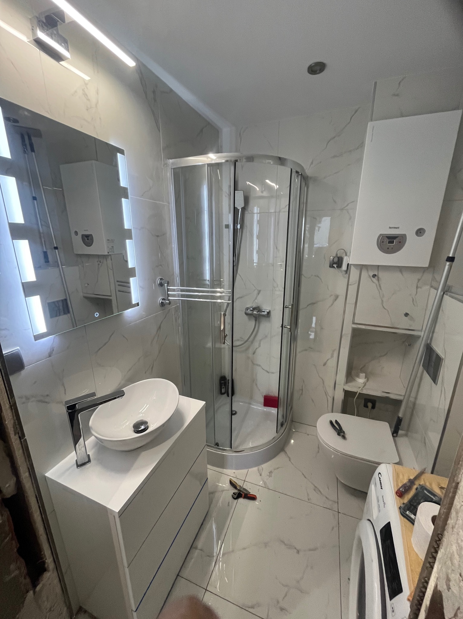 Wykończona łazienka z kabiną prysznicową, umywalką na szafce i toaletą. Białe marmurowe płytki na ścianach i podłodze. Widoczne narzędzia sugerują trwające prace wykończeniowe.