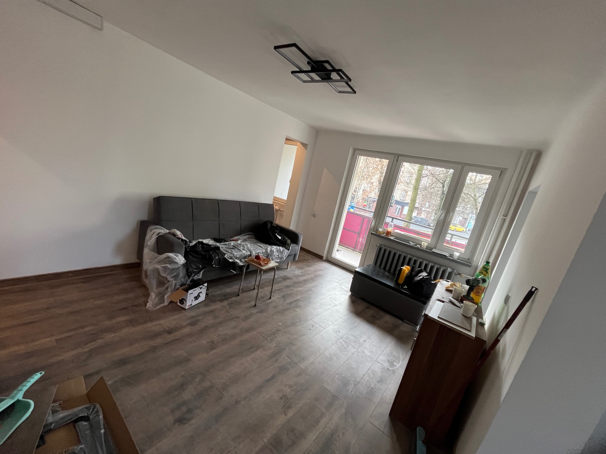 Pokój po remoncie: panele podłogowe, świeżo pomalowane ściany, okno z widokiem na drzewa. Część mebli przykryta folią ochronną. Widoczne narzędzia i akcesoria malarskie.