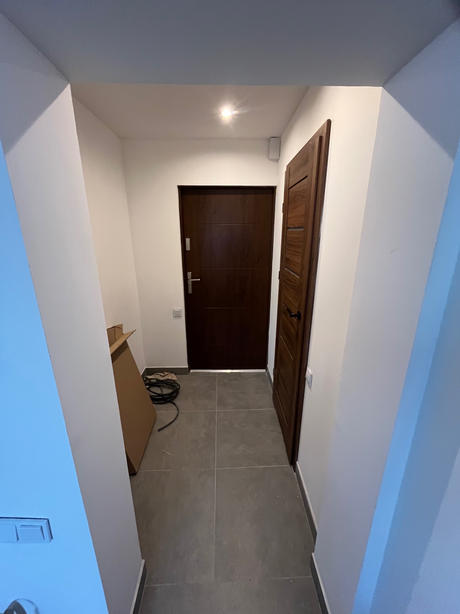 Wnętrze korytarza po remoncie: szare płytki podłogowe, białe ściany, dwoje ciemnych drzwi. Widoczny karton i kable, sugerujące prace wykończeniowe.