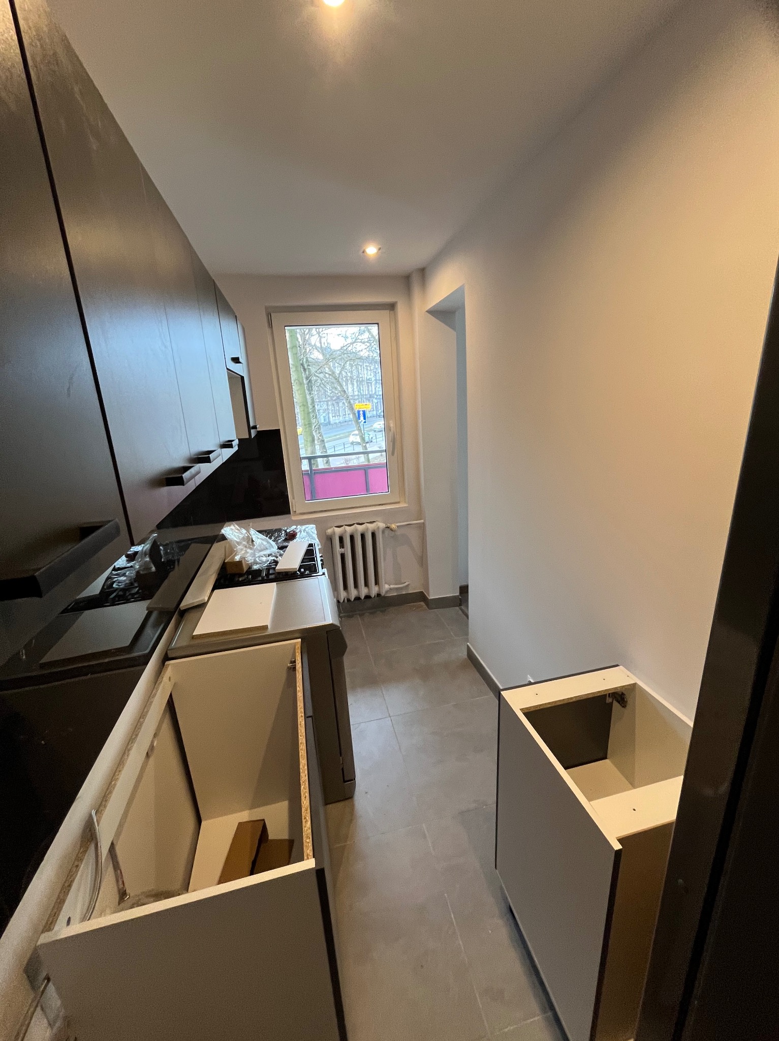 Montaż szafek kuchennych w trakcie remontu. Widoczne niedokończone szafki, okno z widokiem na ulicę i grzejnik. Szare płytki na podłodze.